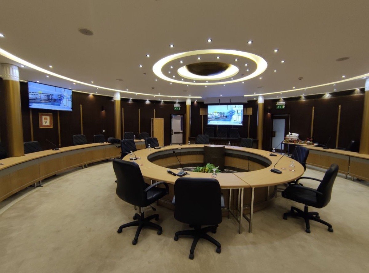 Round meeting table encircling a central planter, with dual wall-mounted displays visible beyond the seating