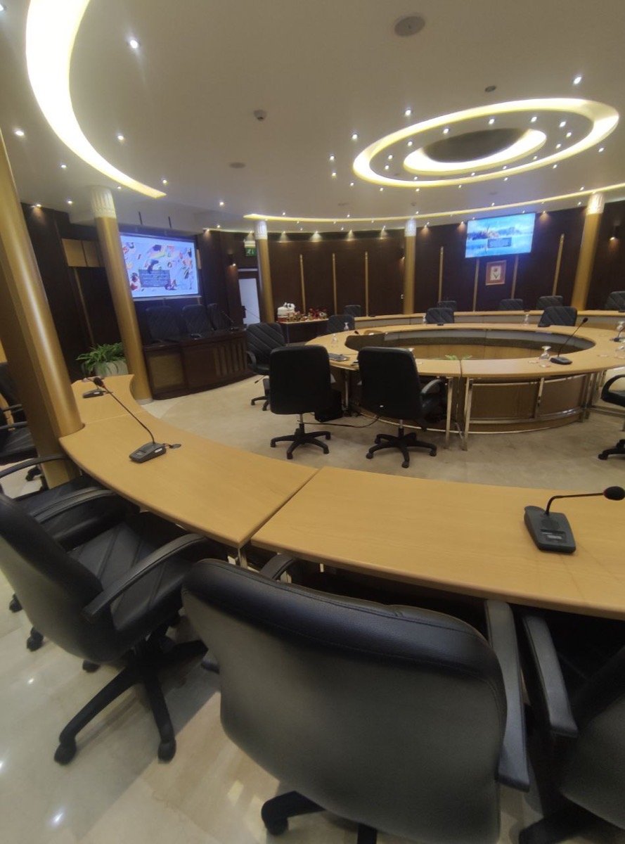 Boardroom seating arranged around the curved table, with a wall-mounted display and decorative lighting column in view
