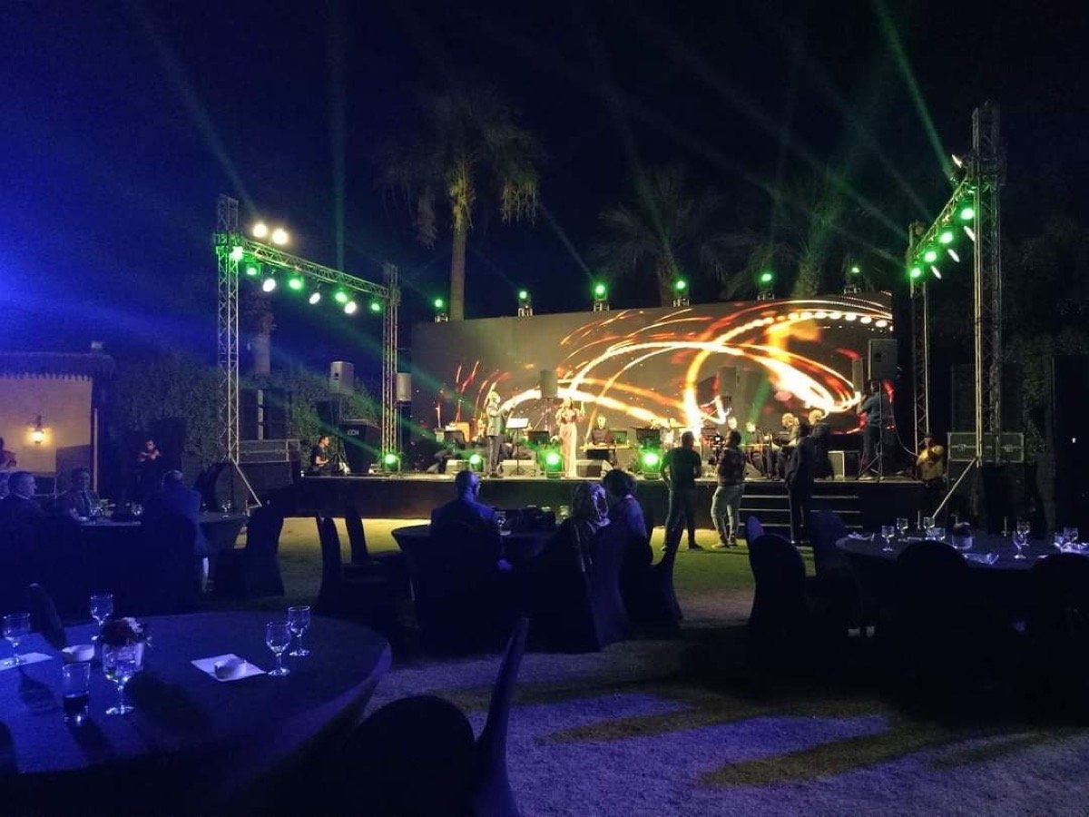 Outdoor evening event with a live band on stage under moving lights and a glowing LED backdrop