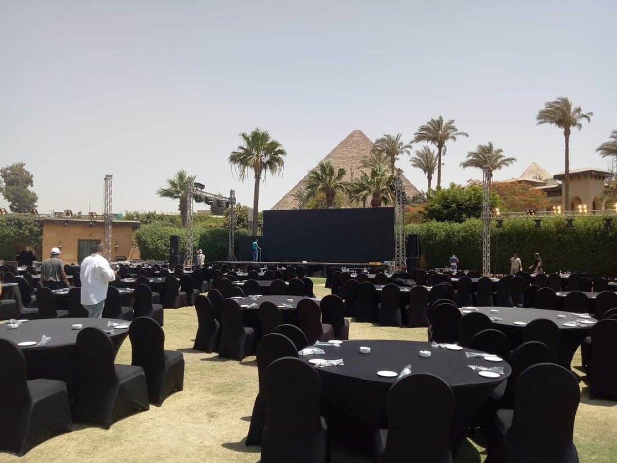 Outdoor daytime event setup with a large LED screen and round tables, the Giza pyramids visible behind